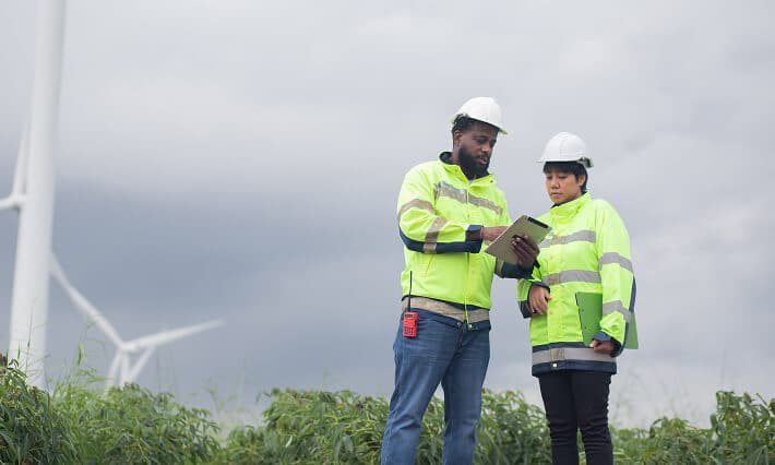 Man and woman wearing hard hats and high visibility jackets stand near windmills looking at a tablet.
