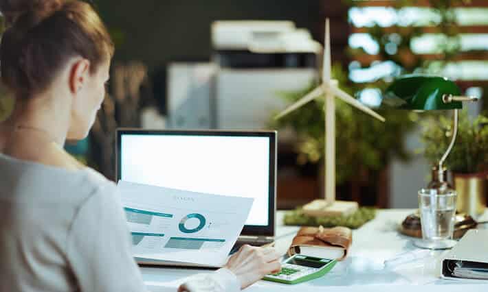 Woman working on a computer while referencing a paper with data.